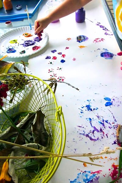 White table cloth with blobs of blue, purple and red paint with a green basket filled with leaves in the foreground