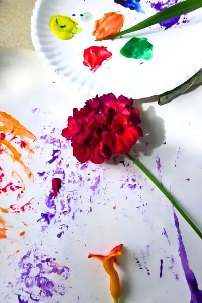 Paper plate with red, green and orange paint blobs next to a red rose with orange and purple paint sprinkles