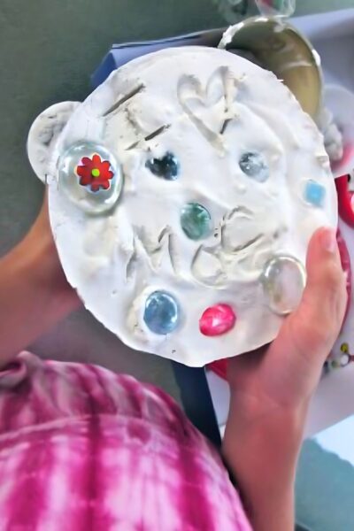 Childs hands holding a white homemade stepping stone with red, blue and clear glass jewels embedded in the stone.