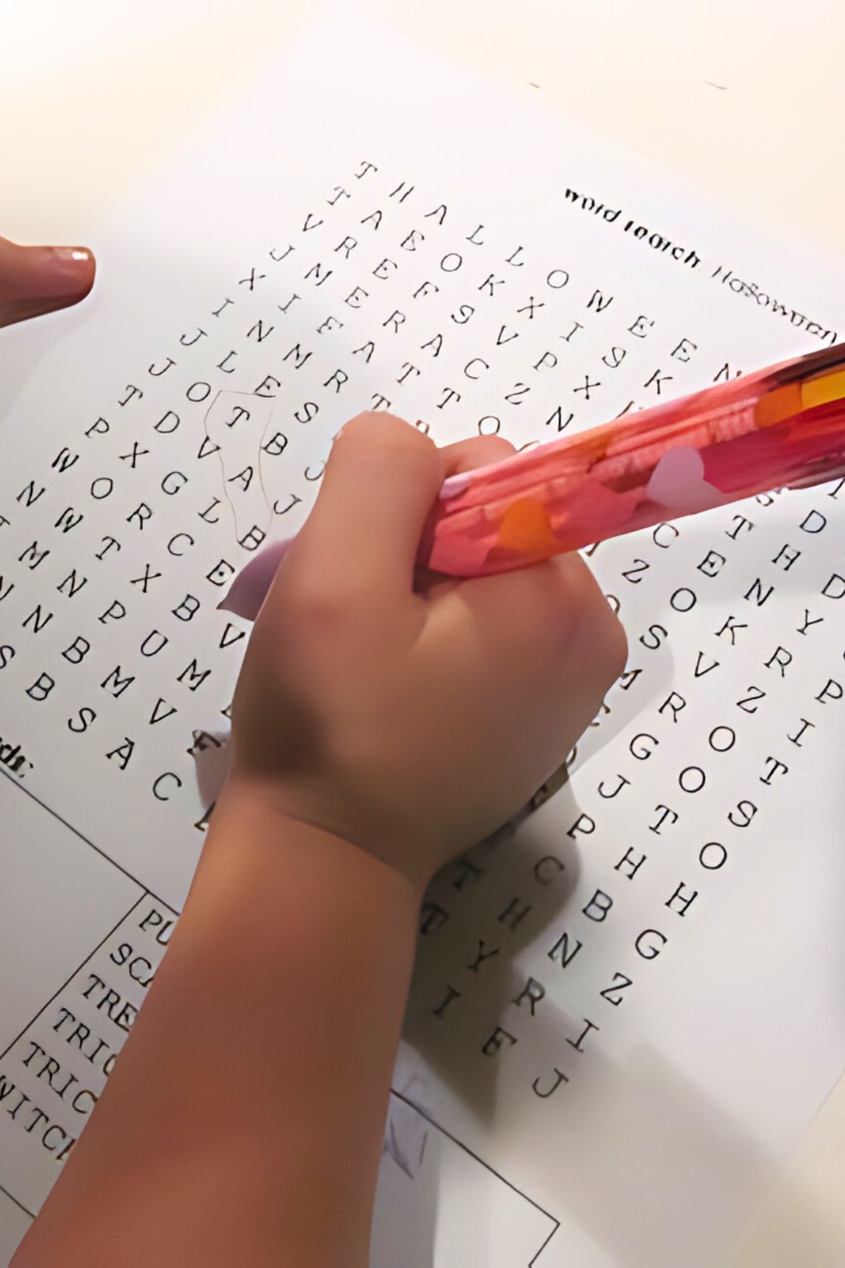 Child's hand holding an orange pen marking a word search