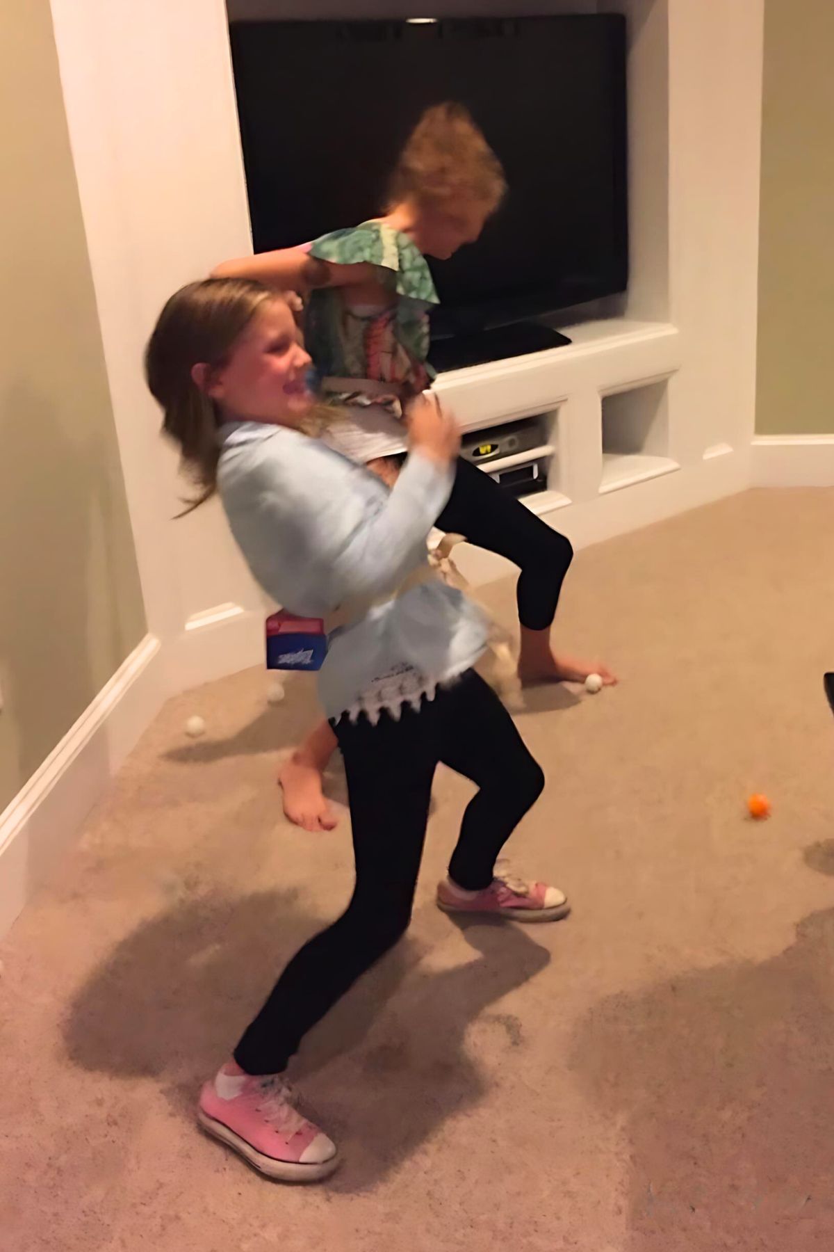 Two girls with a Ziplock baggie box held onto their back by a cloth strap working to bounce ping pong balls onto the floor