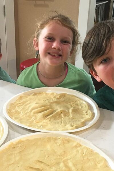 Two young boys and a girl smiling and showing off homemade pizza crust with their handprints in the center