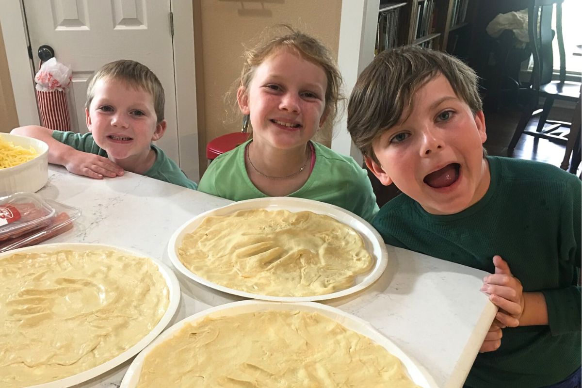 Two young boys and a girl smiling showing off homemade pizza crusts with their handprints in the center