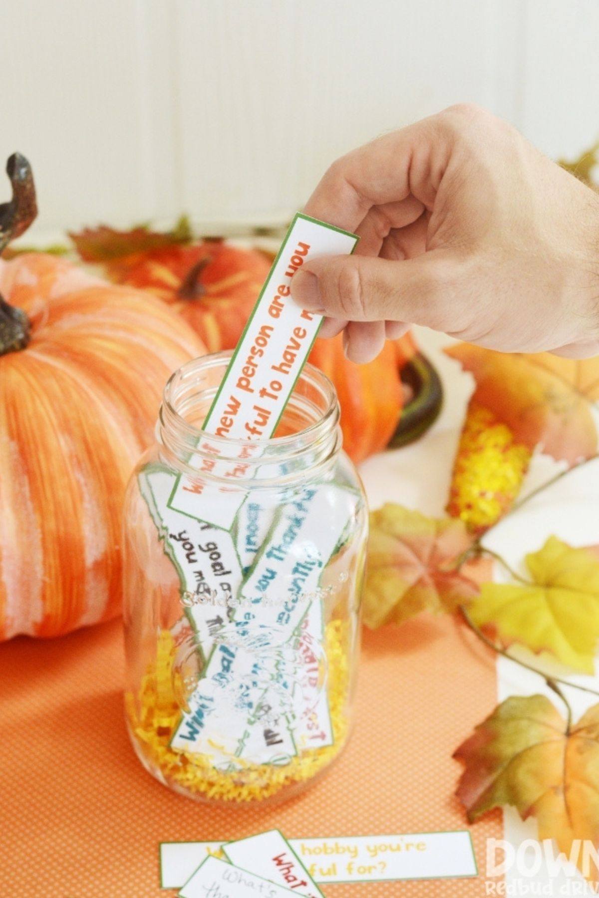 Paper slips printed with gratitude questions in a mason jar. A hand is pulling one slip from the jar