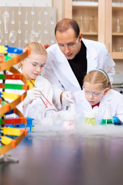 A man and two girls, all dressed in white lab coats, with beakers on a table