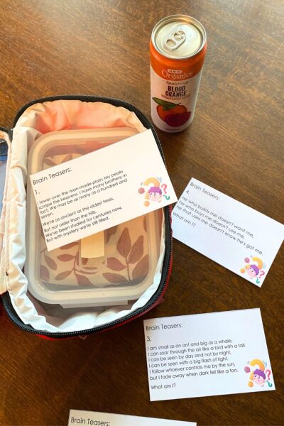 Red soft sided lunch pail with an orange drink and brain teaser lunchbox notes on a wooden table