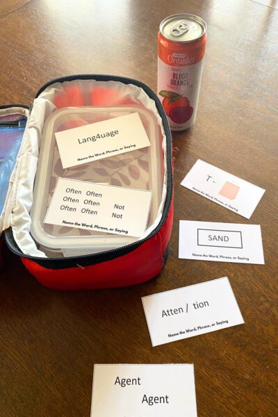 Red soft side lunch pail and orange drink on a dark wood table with printed rebus word puzzle cards