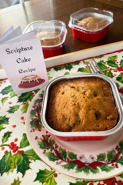 Brown cake with raisins in a square red foil tin on a Christmas plate and fork with two more tins behind on a wooden table