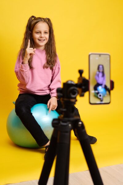 A young girl in a pink sweater sits on a blue rubber ball in front of a yellow wall with a cell phone on a tripod recording
