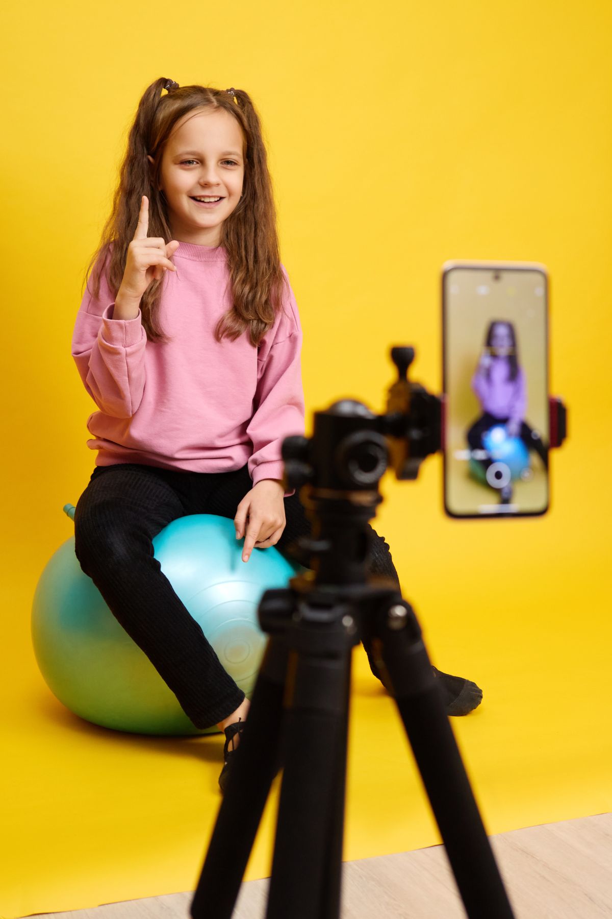A young girl in a pink sweater sits on a blue rubber ball in front of a yellow wall with a cell phone on a tripod recording