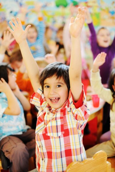 Elementary-aged kids in a classroom with their hands raised and laughing