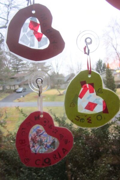 Kid-made paper hearts with a clear contact paper pane in the center decorated with stamps and bits of colored paper hanging in front of a window