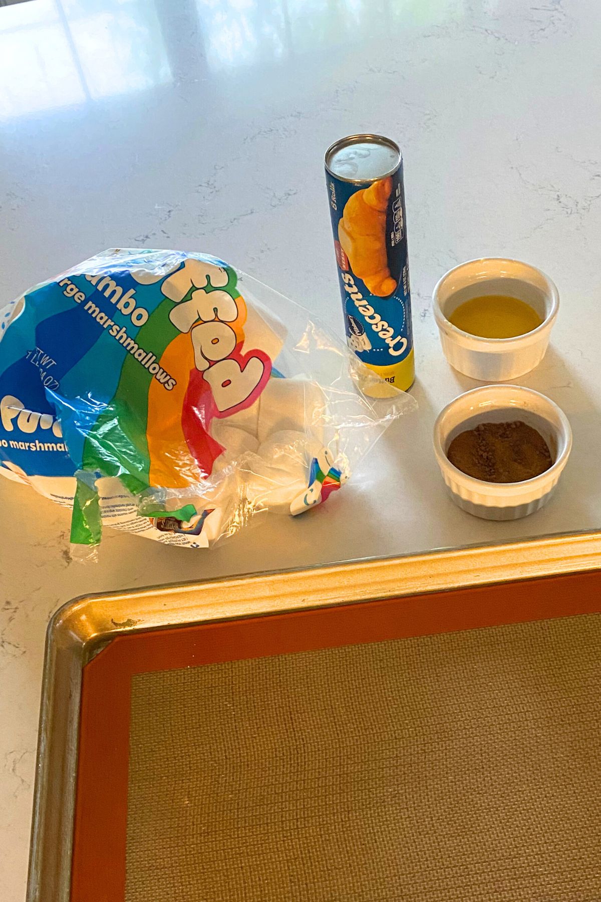 Glass containers of brown sugar and butter, Crescent Roll package, marshmallows and a cookie sheet on a white counter