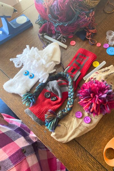Three sock puppets on a wooden table with thread and buttons scattered around them