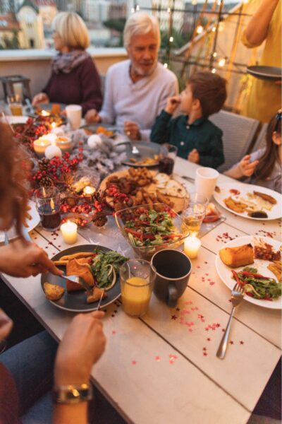 Family seated outdoors at a wooden table with red berry decorations, dinner set, candles and soft lights around them.