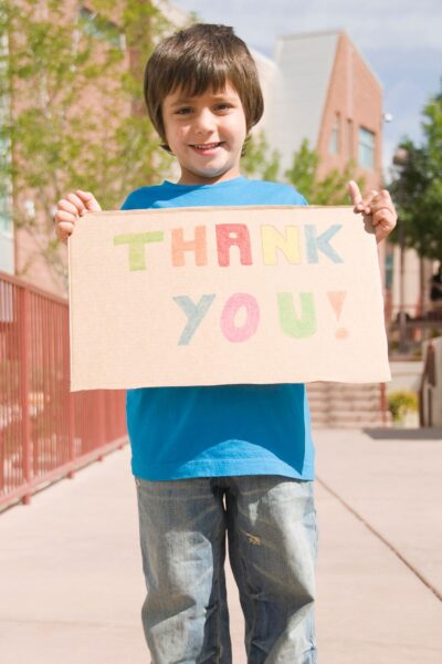 A young boy wearing a blue shirt and holding a handmade cardboard thank you sign.