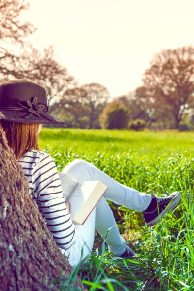 A girl wearing a hat and reading a book under a tree in a grassy field